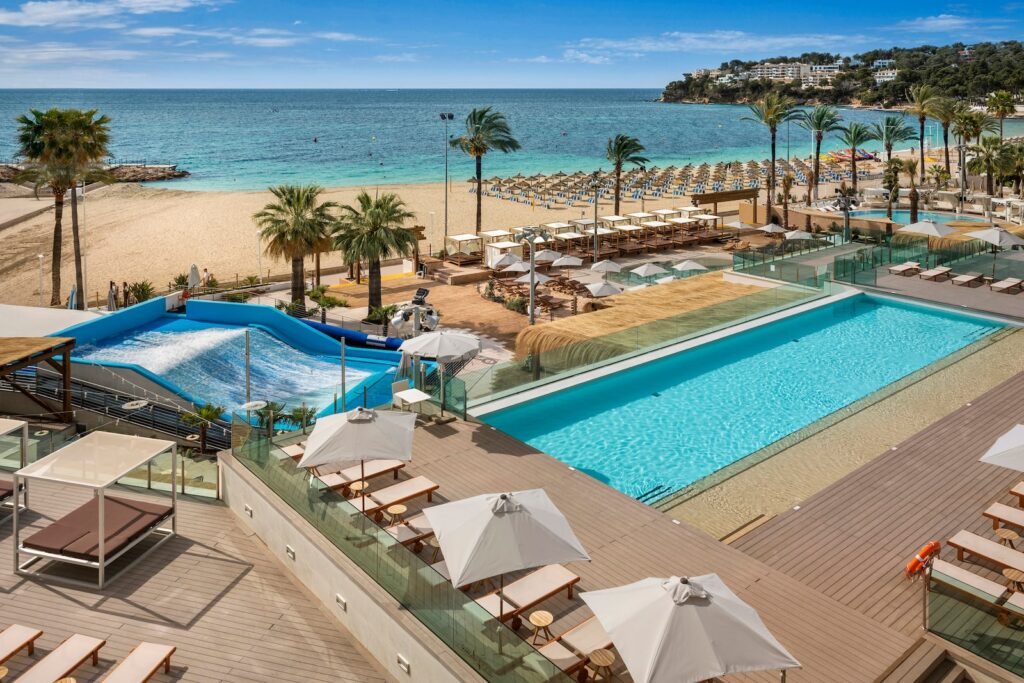 A beachfront resort features a large pool, sun loungers with umbrellas, palm trees, a wave simulator, and direct access to a sandy beach with rows of lounge chairs overlooking the turquoise sea under a blue sky.
