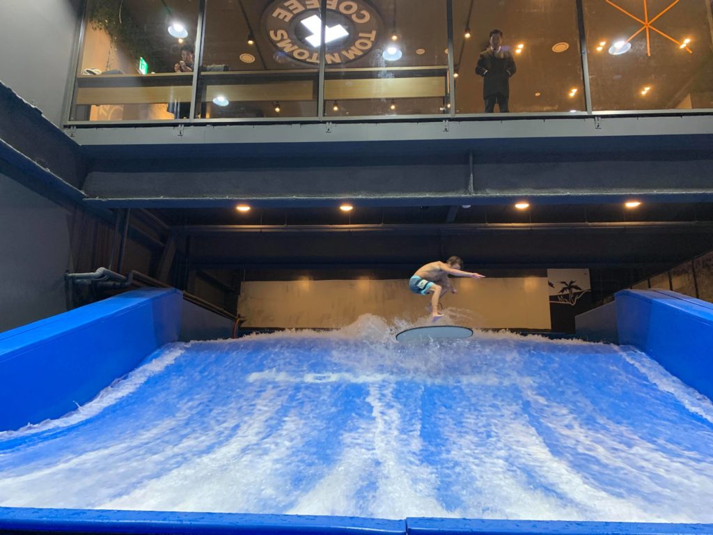 A person wearing swim trunks is surfing on an indoor artificial wave under a balcony with people watching from behind glass at a café called Tom N Toms Coffee.