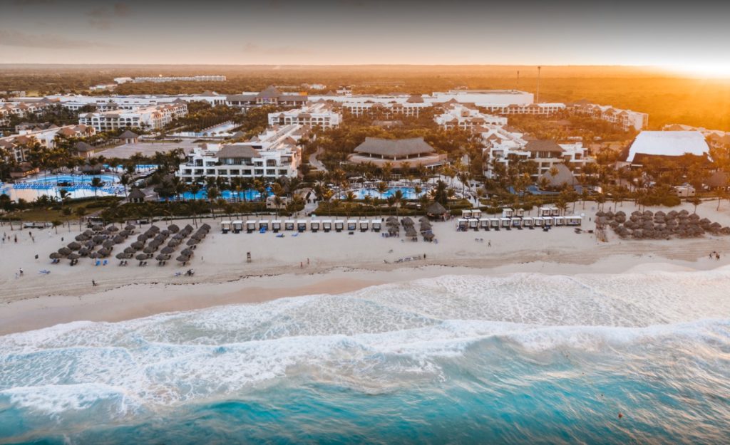 Aerial view of a beachfront resort at sunset, showing white sand, rows of loungers and umbrellas, turquoise waves, multiple pools, lush greenery, and large hotel buildings in the background.
