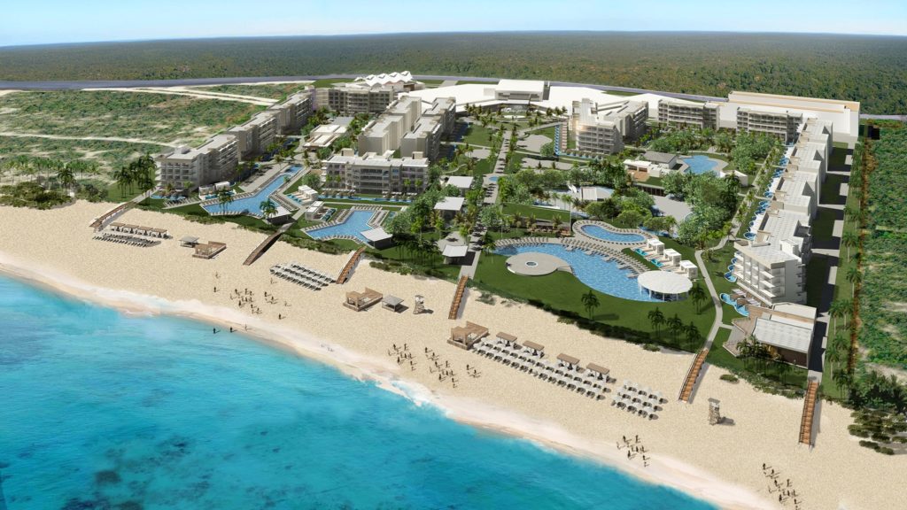 Aerial view of a large beachfront resort with multiple pools, palm trees, lounge areas, and walkways, set along a sandy beach with clear blue water and sun loungers arranged by the shore.