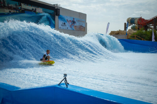A person rides a yellow bodyboard on the FlowBarrel Double, catching artificial blue waves at a water park. A large wave bends behind them, water slides stand in the background, and a camera is set up on a tripod by the pool’s edge.