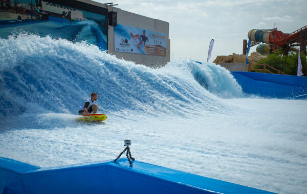 A person rides a yellow bodyboard on the FlowBarrel Double, catching artificial blue waves at a water park. A large wave bends behind them, water slides stand in the background, and a camera is set up on a tripod by the pool’s edge.
