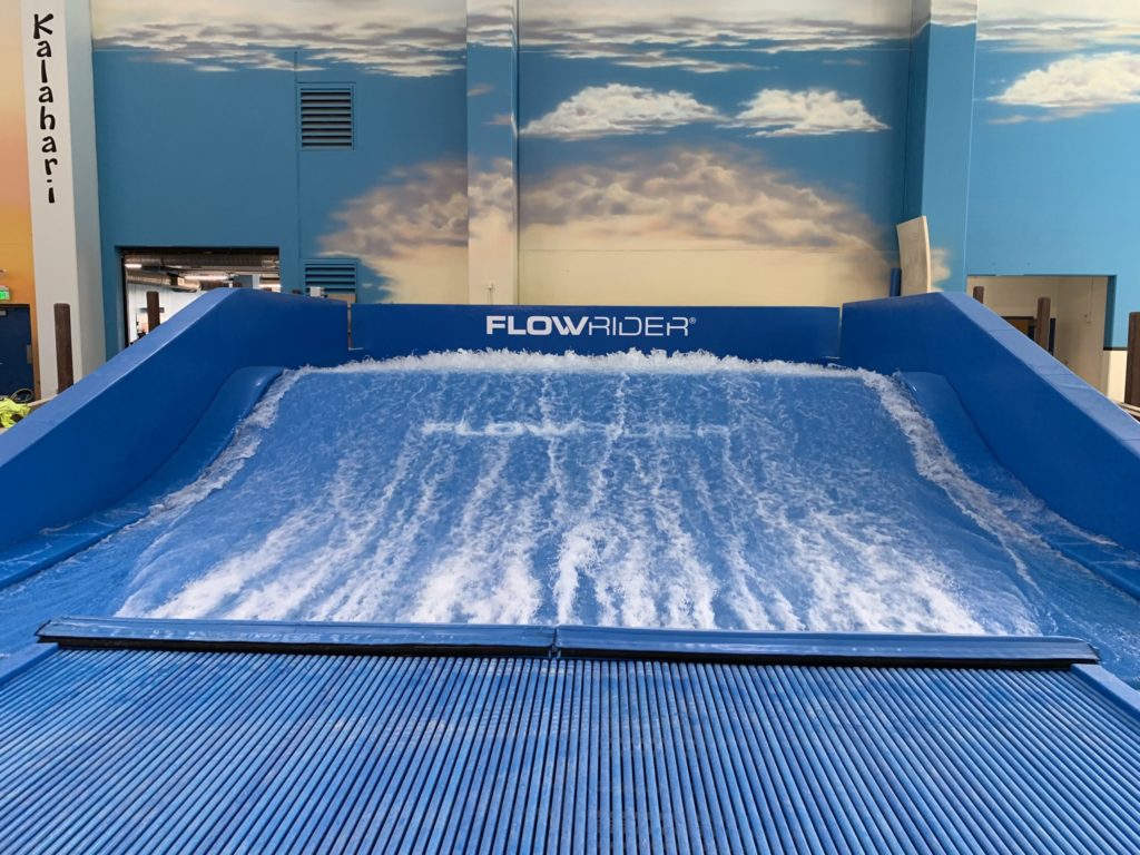 A blue indoor FlowRider surfing simulator with artificial waves and FLOWRIDER written on the back wall; sky and clouds are painted on the surrounding walls.