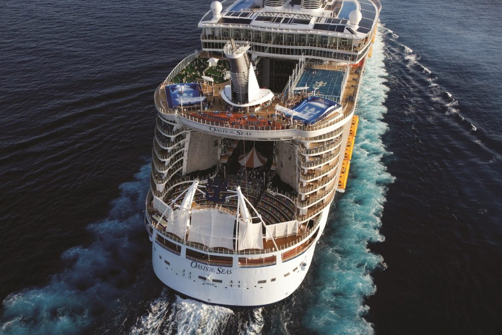 Aerial view of the cruise ship Oasis of the Seas sailing on the ocean, with blue wake behind and multiple decks visible, including outdoor pools and recreational areas.