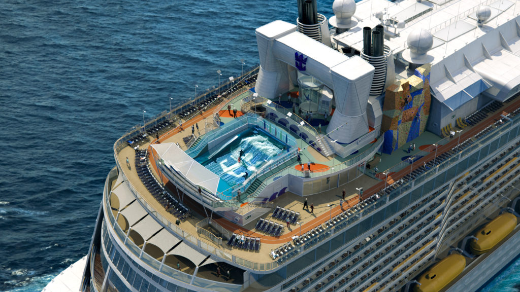 Aerial view of the top deck of a cruise ship at sea, featuring a surfing simulator with people riding artificial waves, lounge chairs, and observation areas.