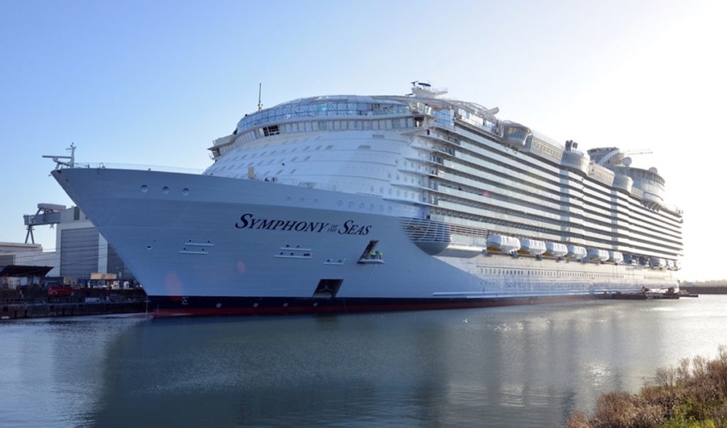 A large white cruise ship named Symphony of the Seas docked at a port, reflected in calm water with blue sky in the background.