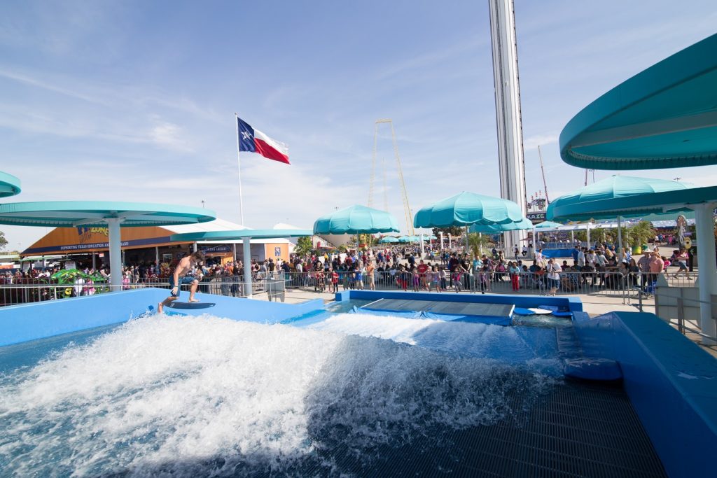 A person surfs on an artificial wave at an outdoor water park attraction, with a crowd watching behind a fence. A large Texas flag and several turquoise umbrellas are visible under a sunny sky.