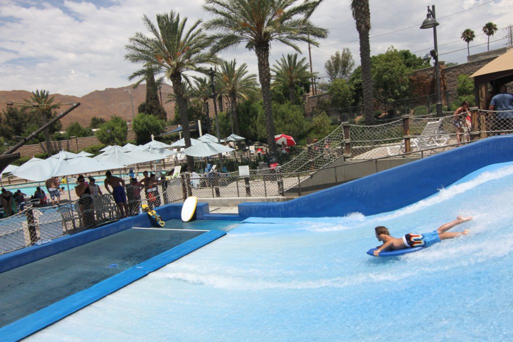 A person rides a bodyboard on a blue artificial wave at a water park, with people watching nearby, palm trees, umbrellas, and mountains in the background.