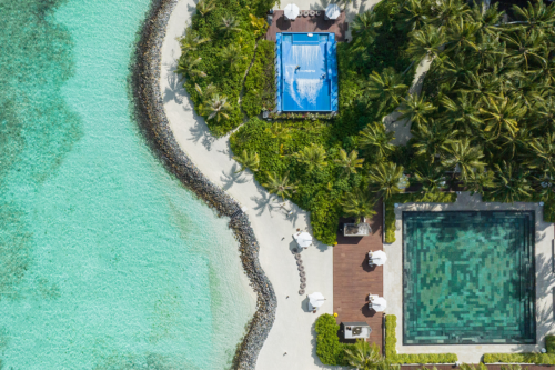 Aerial view of a tropical resort with turquoise ocean, sandy beach, lush palm trees, a rectangular mosaic-tiled pool, a FlowRider Wave in a Box for surfing fun, and white umbrellas on a wooden deck.