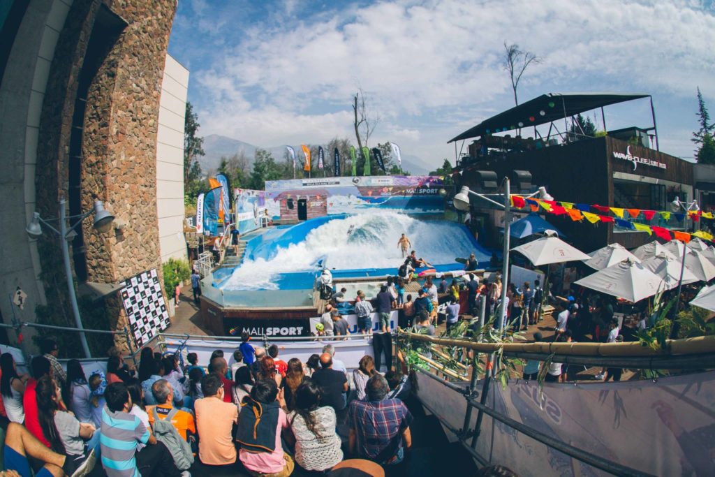 A crowd watches an indoor surfing competition at a wave pool, with surfers riding artificial waves while spectators sit and stand around the pool under colorful flags and umbrellas.
