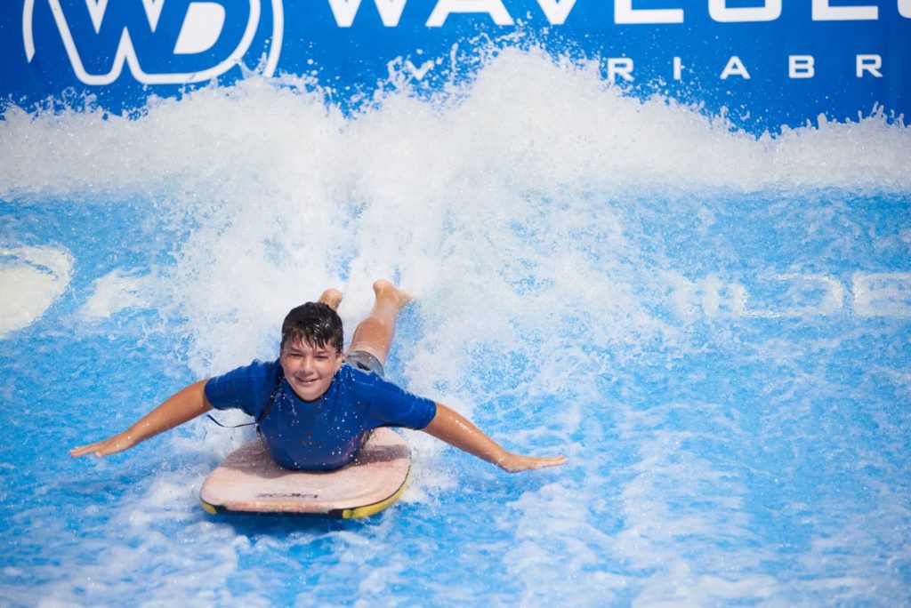 A smiling boy in a blue shirt lies on a surfboard, riding artificial waves at an indoor surfing facility, with water splashing around him.