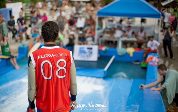 A person wearing a red FLOWRIDER 08 jersey stands at the edge of a flowrider surf simulator, facing a crowd of spectators and judges under a blue tent at an outdoor event.