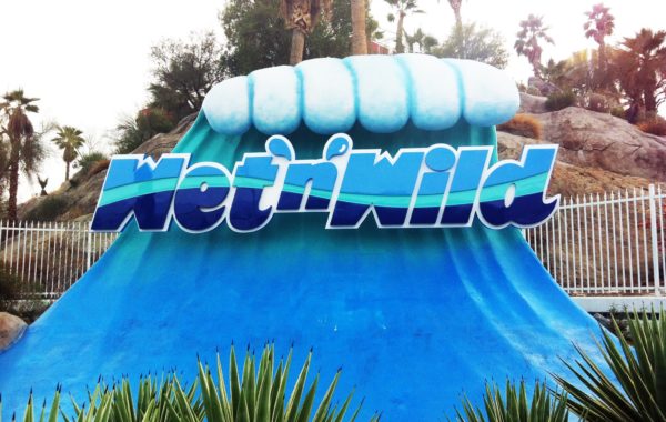 A large blue wave sculpture with the words WetnWild in bold letters stands in front of palm trees and rocky terrain, surrounded by green plants and a white fence in the background.