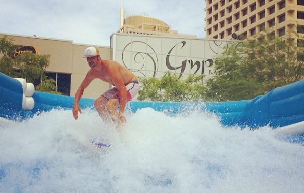A man wearing swim trunks, sunglasses, and a cap is surfing on an artificial wave at an outdoor urban location, with buildings and hotel signage visible in the background.