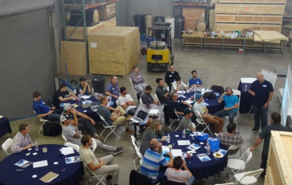 A group of people sit at round tables in a warehouse, listening to a man standing and speaking near a screen. Laptops, papers, and drinks are visible on the tables. Large crates and industrial equipment are in the background.