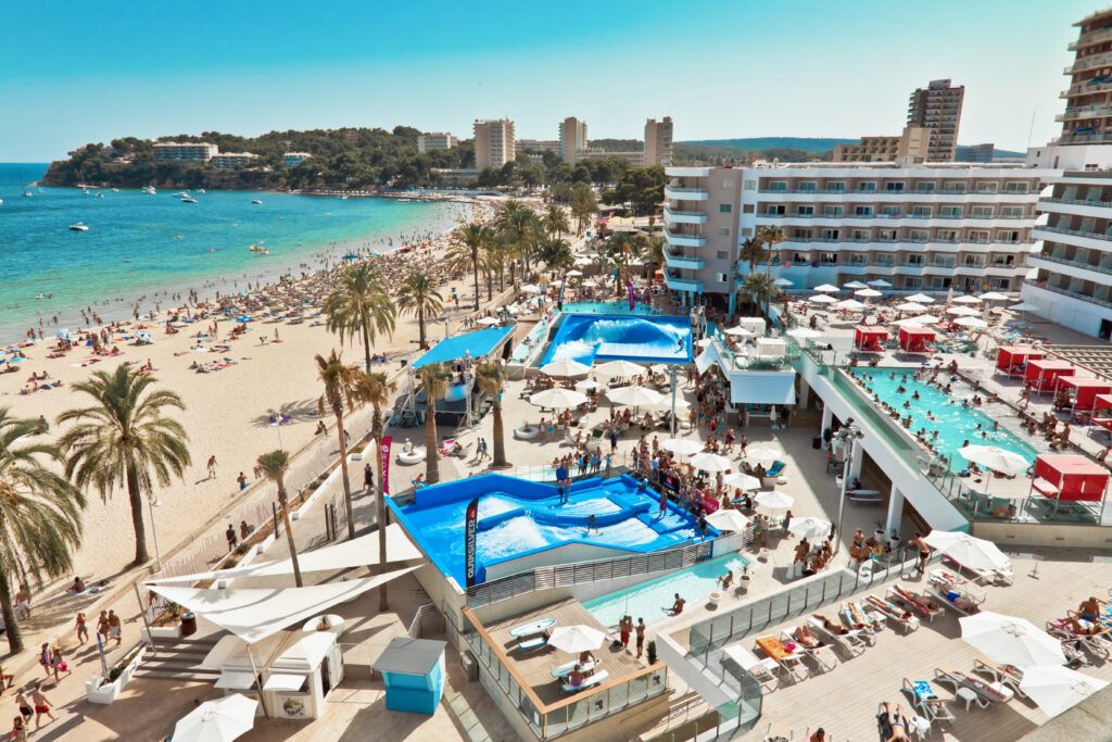 A lively beach resort with palm trees, a crowded sandy beach, turquoise sea, and a pool area with sun loungers, people swimming, and a wave simulator. Modern hotels and tall buildings line the shore under a clear blue sky.