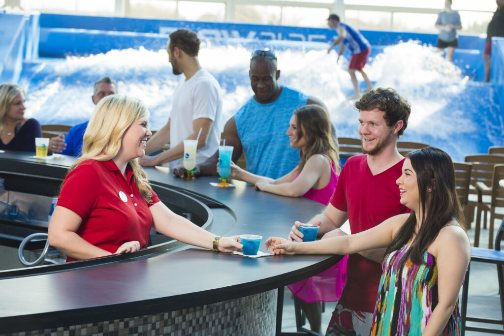 A smiling bartender in a red shirt serves drinks to a man and woman at an indoor bar, with other people seated nearby and a person surfing on an indoor wave machine in the background.