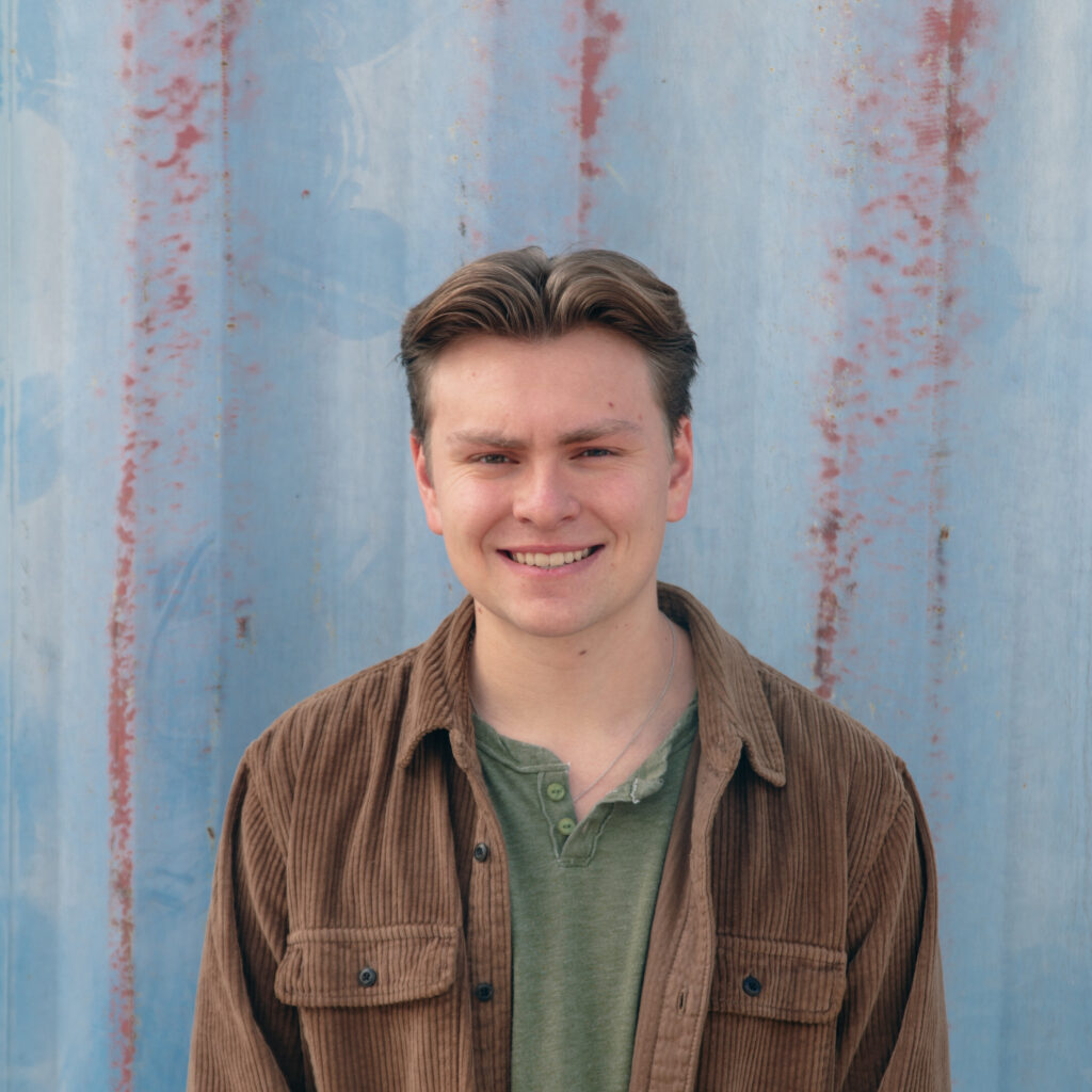 A young man with short brown hair, wearing a green shirt and a brown corduroy jacket, smiles while standing in front of a weathered, light blue wall with vertical rusty streaks.
