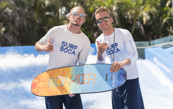 Two men wearing sunglasses and Surf Boca shirts stand on a wave simulator, smiling and giving thumbs up. One holds a colorful FlowRider board. Palm trees and blue water are visible in the background.