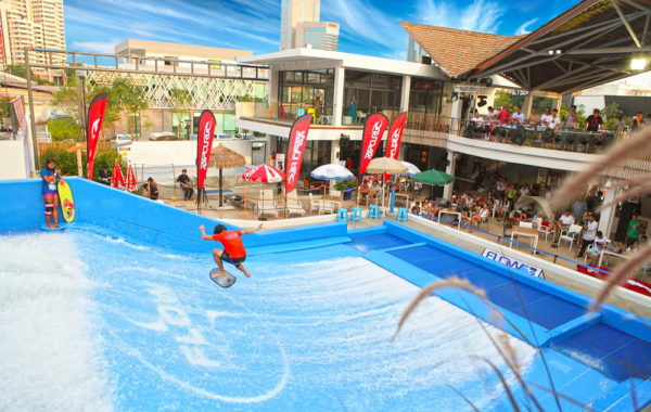 A person in an orange shirt surfs on an artificial wave at an urban outdoor surf park, surrounded by people watching from nearby seating areas and under umbrellas, with city buildings in the background.