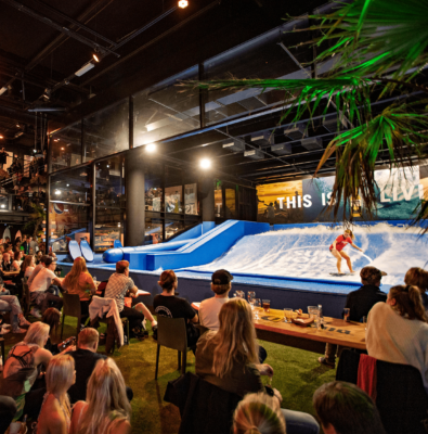 Indoor venue with people seated at tables and watching a person surfing on an artificial wave machine. The atmosphere is lively, with warm lighting and palm plants adding to the beachy vibe.