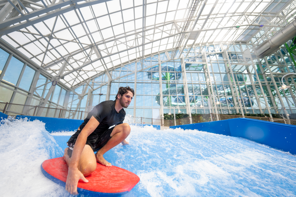 A man wearing a black rash guard rides a red board on an indoor wave simulator, surrounded by a bright, modern glass structure with sunlight streaming through the ceiling.