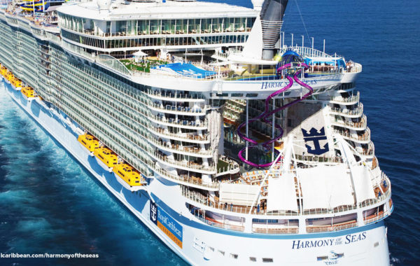 A large white cruise ship, Harmony of the Seas, sails on blue ocean water. The ship features yellow lifeboats, outdoor decks, pools, and a spiraling purple waterslide at the rear.