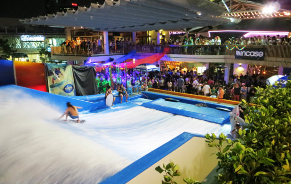 Indoor surfing attraction at night with two people riding artificial waves, surrounded by a large crowd of spectators, colorful lights, and a lively atmosphere at a multi-level venue.