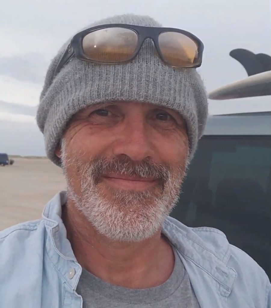A man with a gray beard and mustache smiles at the camera. He wears a gray knit beanie with sunglasses on top and a light blue jacket over a gray shirt. The background shows a beach and a parked vehicle.