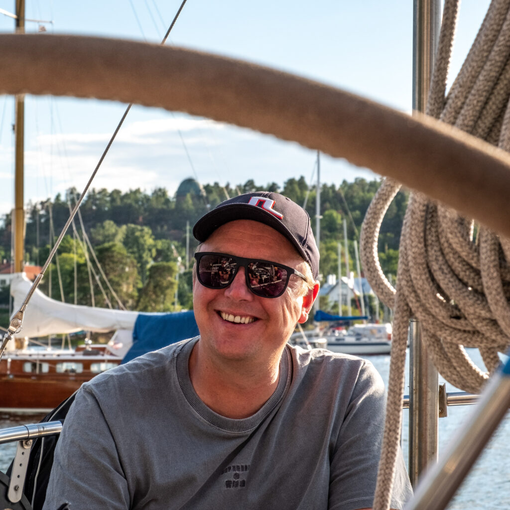 A man wearing sunglasses, a black cap, and a grey t-shirt smiles while sitting on a sailboat. Coiled ropes and boats are visible in the background on a sunny day.