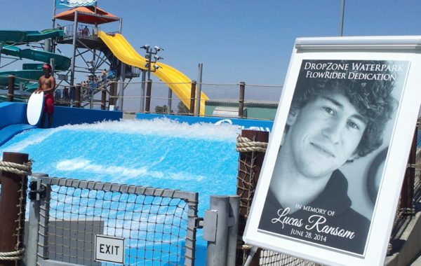 A waterpark wave machine is in use with a lifeguard nearby. In the foreground, a sign displays a black-and-white photo of a young man and reads “DropZone Waterpark Flowrider Dedication in memory of Lucas Ransom, June 28, 2014.”.