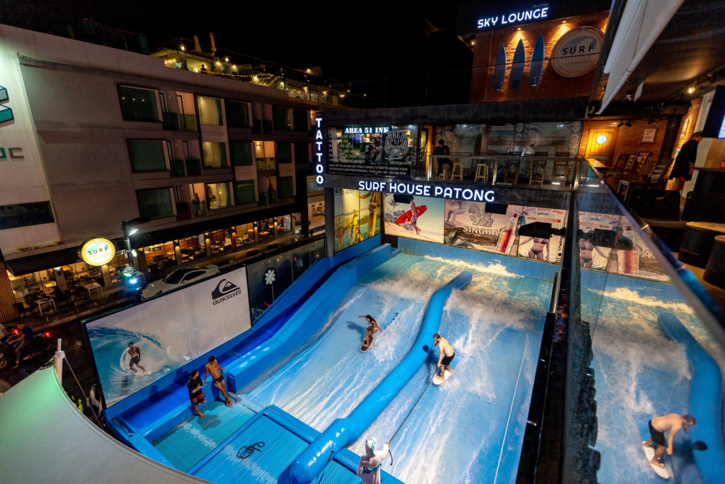 People surf on artificial wave machines at SUP House Patong, an indoor surf venue surrounded by buildings with neon signs at night. Spectators watch from nearby platforms and balconies.