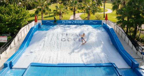 A person surfs on an artificial wave at a FlowRider attraction, surrounded by palm trees and a park-like setting under a sunny sky. Another person stands at the top, preparing to surf.