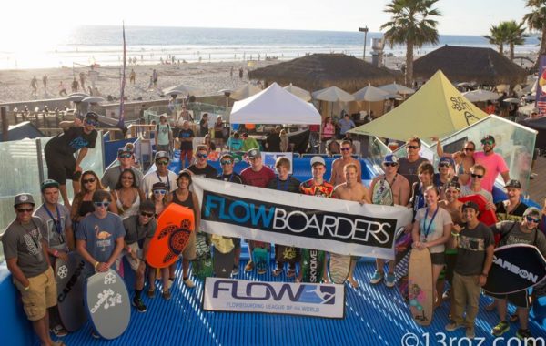 A large group of people holding flowboards and a “FLOWBOARDERS” banner poses for a photo at a sunny beach event, with palm trees, umbrellas, and the ocean in the background.
