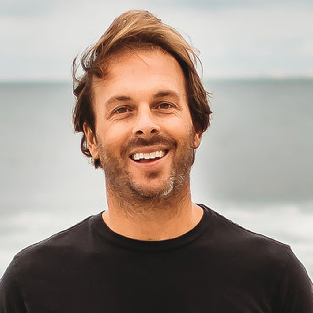 A man with tousled brown hair and facial stubble smiles at the camera while standing outdoors near the ocean, wearing a black t-shirt, with a cloudy sky and water in the background.