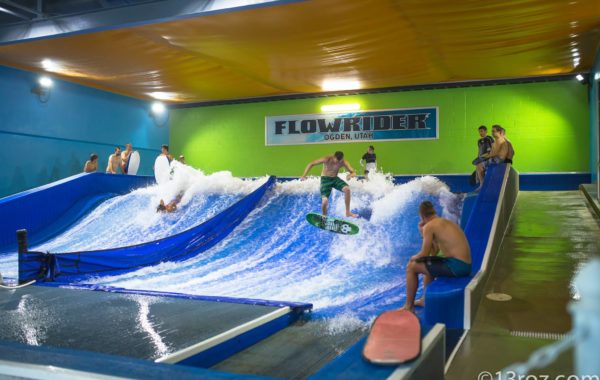 Several people surf and watch on an indoor artificial wave machine under a yellow ceiling. One surfer rides a wave, while others wait or sit on the side. A FlowRider sign is visible on the green wall in the background.