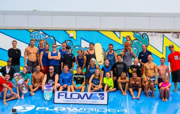 A large group of people of various ages in swimwear and wetsuits pose and smile on a blue artificial wave ride platform, with surfboards and a colorful mural in the background.