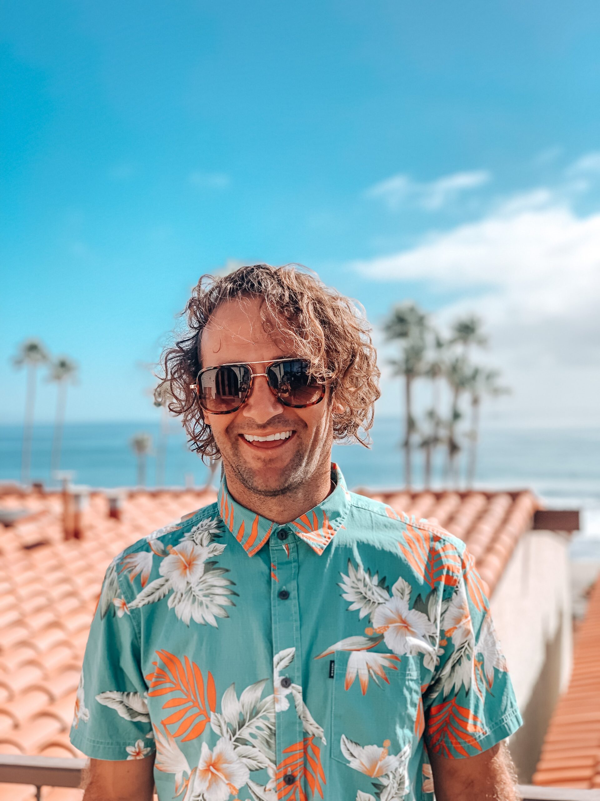 A man wearing sunglasses and a blue floral shirt stands smiling in front of a tiled rooftop, with palm trees and the ocean in the background under a bright, blue sky.
