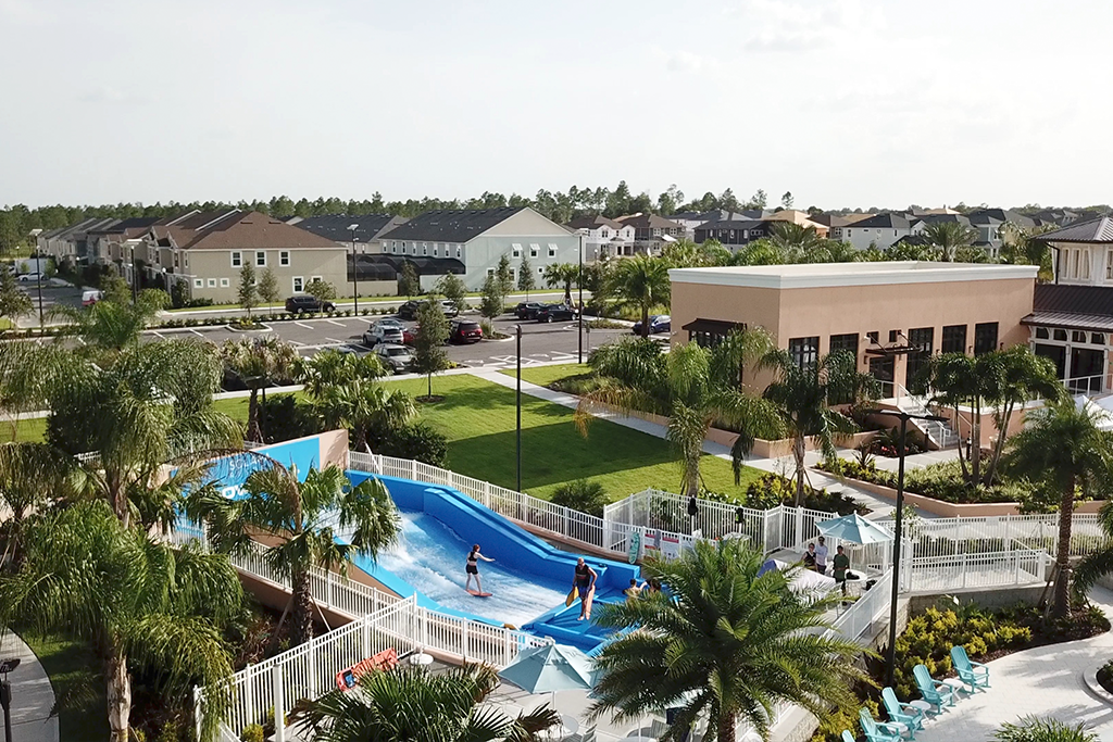 Aerial view of a residential community with a wave pool and people surfing, surrounded by palm trees, a parking lot, and modern apartment buildings in the background.