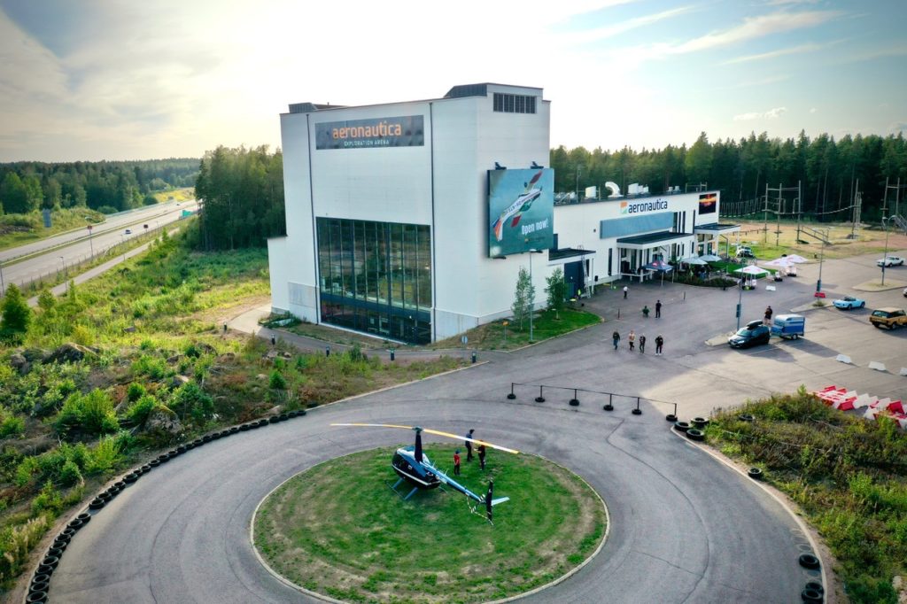 Aerial view of a modern Aeronautica building surrounded by trees, with a helicopter parked on a circular landing pad and several people and cars nearby, next to a highway under a clear sky.