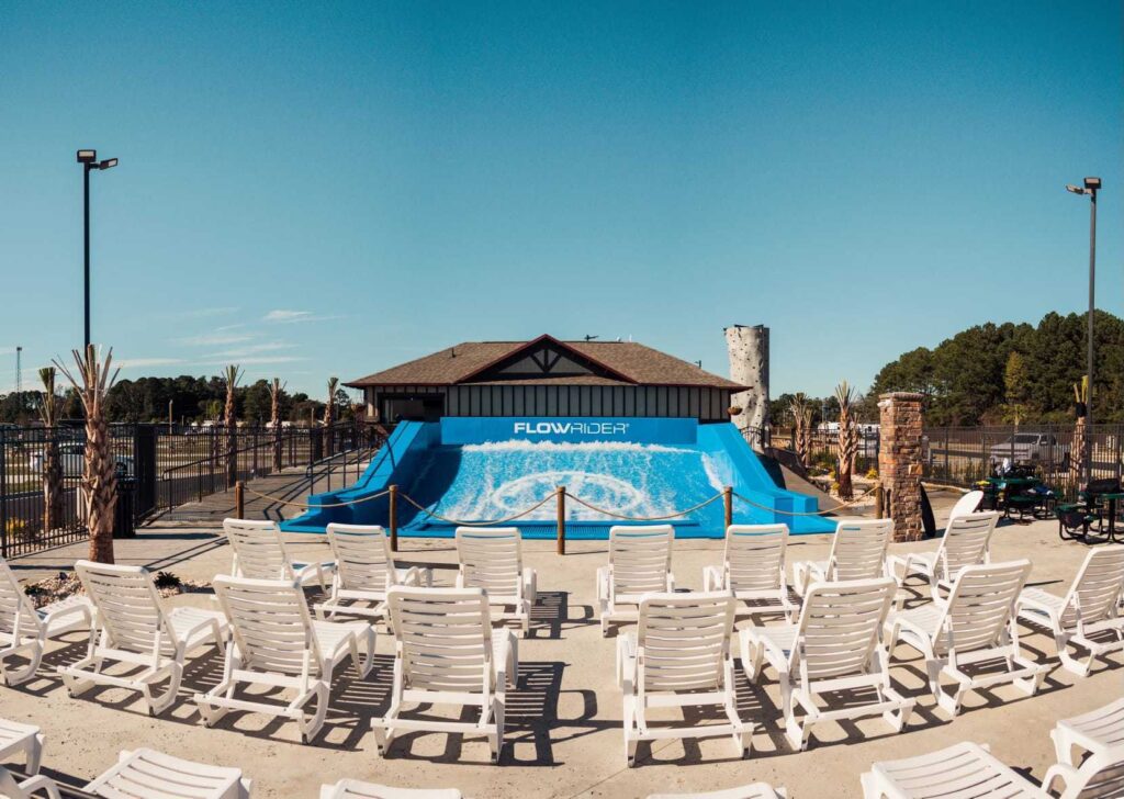 Rows of empty white lounge chairs face a blue FlowRider surf simulator under a clear sky, with trees and a building in the background at an outdoor recreation area.