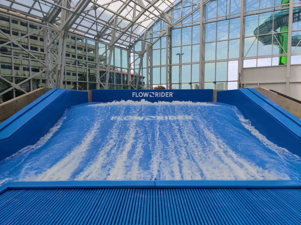 A blue indoor FlowRider surf simulator generates artificial waves inside a glass-walled building, with foam and water flowing upward toward the viewer.
