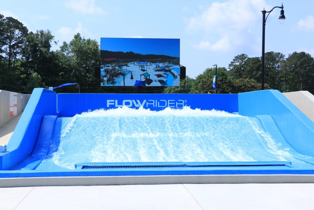 A FlowRider wave machine generates artificial waves for surfing in an outdoor setting, with a blue ramp, trees in the background, and a large screen displaying a water activity scene.