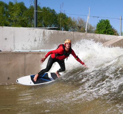 A person wearing a helmet and wetsuit surfs on an artificial wave in a concrete water channel, leaning forward as water splashes behind them under a clear blue sky.
