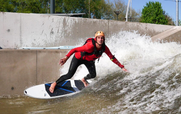 A person wearing a helmet and wetsuit surfs on an artificial wave in a concrete water channel, leaning forward as water splashes behind them under a clear blue sky.