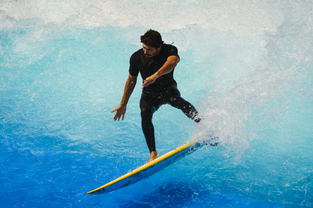 A man in a black wetsuit surfs on a yellow FlowSurf board, balancing skillfully as water splashes around him against a backdrop of blue ocean waves.