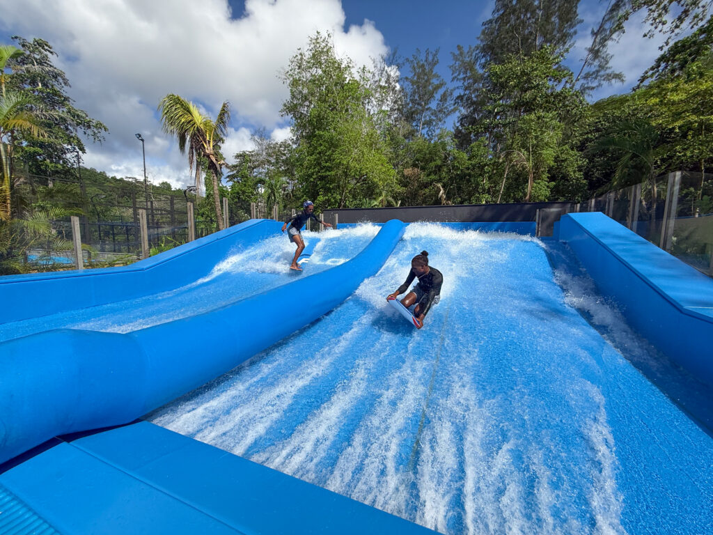 Two people ride artificial surf waves on blue water slides surrounded by lush green trees under a partly cloudy sky. One person is upright while the other is crouched low on a board.