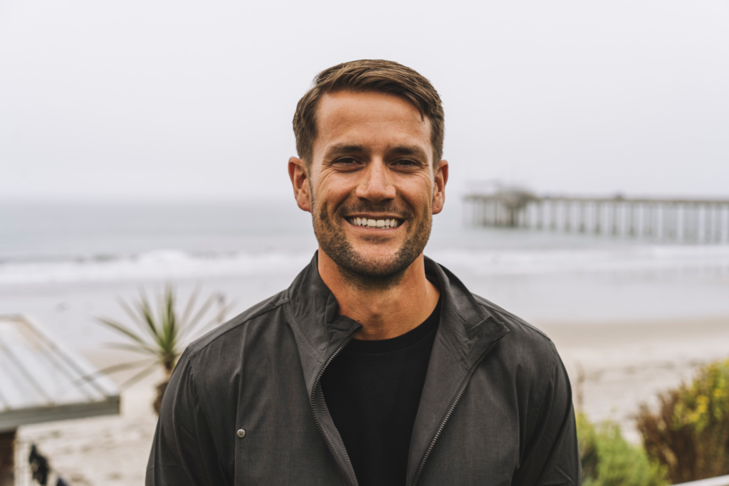 A man with short brown hair and a beard smiles while standing on a beach. He is wearing a dark jacket and a black shirt. The ocean, sandy shore, plants, and a pier are visible in the background under a cloudy sky.