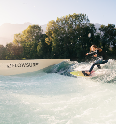 A person in a wetsuit surfs on an artificial wave at a facility with FLOWSURF branding, surrounded by trees, under a clear sky with sunlight streaming through.