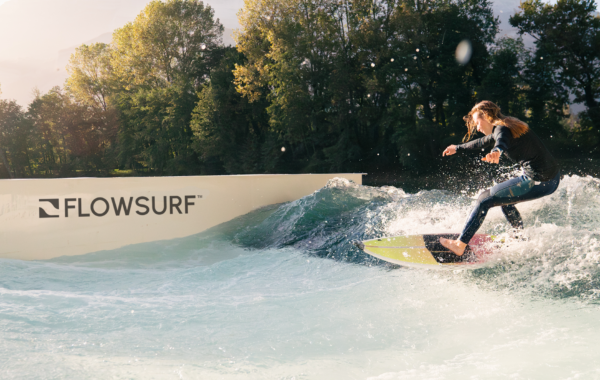A person in a wetsuit surfs on an artificial wave at a facility with FLOWSURF branding, surrounded by trees, under a clear sky with sunlight streaming through.